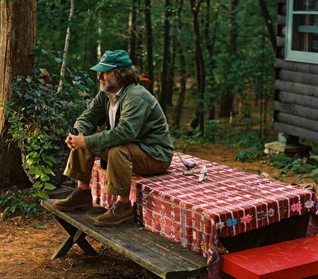 Man sitting on a wooden bench by a cabin in the woods with a red checkered tablecloth.